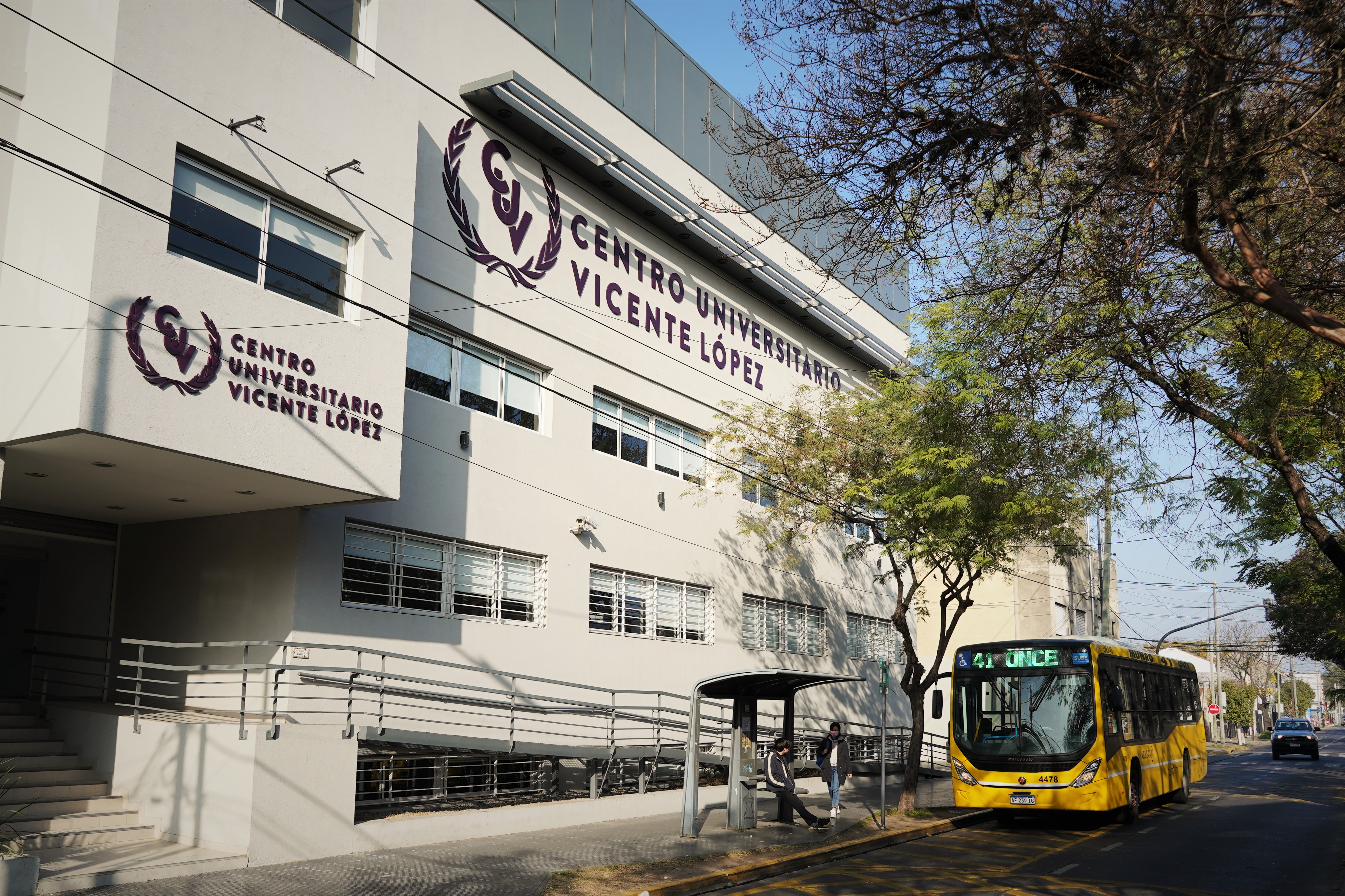 Fachada del edificio del Centro Universitario Vicente López en Munro, con un autobús amarillo estacionado en la parada frente al centro.