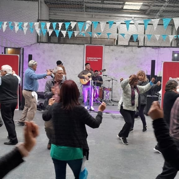 Un grupo de personas disfrutando de una tarde de música y baile en la Sociedad de Fomento de Villa La Ñata durante el ciclo Domingos de Peña, con una banda tocando en el fondo.