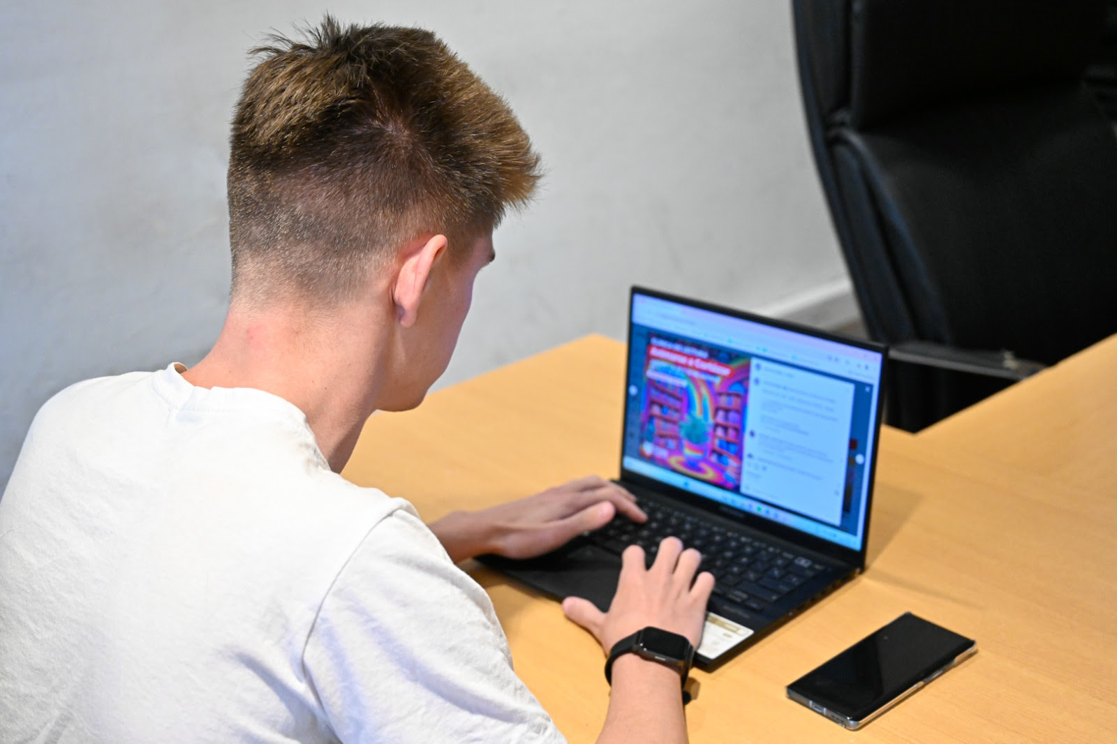 Una persona con una camiseta blanca, de espaldas a la cámara, utiliza una computadora portátil en un escritorio. En la pantalla, se observa la información sobre el evento "Animarse a Cortázar". Al lado de la computadora, hay un teléfono móvil negro sobre la mesa.