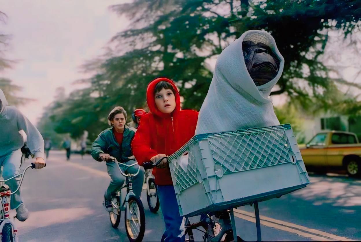 Escena icónica de la película "E.T. El Extraterrestre", dirigida por Steven Spielberg, donde un niño con una sudadera roja monta una bicicleta con una cesta en la que se encuentra E.T. envuelto en una manta blanca, seguido por otros niños en bicicletas. Esta imagen es parte de la programación de cine de aventuras del Centro Cultural Munro en Vicente López.