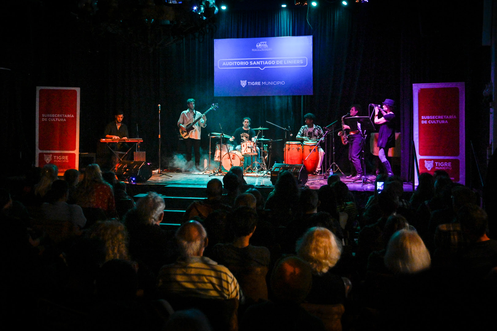 Banda de jazz tocando en el escenario del auditorio del Museo de la Reconquista, con el público sentado disfrutando del espectáculo. Banners de la Subsecretaría de Cultura del Municipio de Tigre en los laterales del escenario.