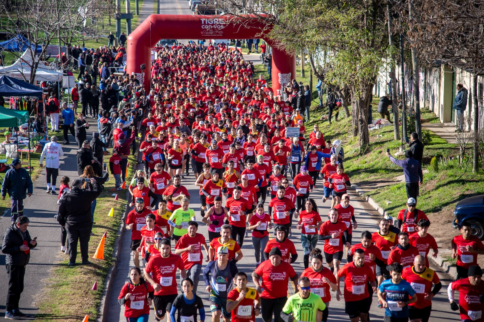 Gran cantidad de corredores participando en la Carrera Comunidad de Tigre, todos con camisetas rojas, al inicio de la carrera bajo un arco inflable rojo en El Talar.