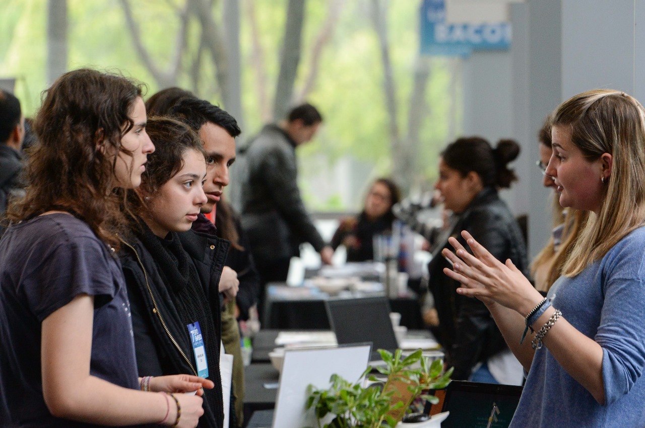 Grupo de jóvenes hablando con una expositora en uno de los stands de Expo Joven, en un ambiente de feria con diversas personas y actividades en segundo plano.