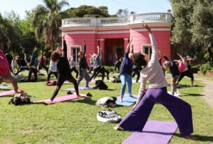 Grupo de personas realizando una clase de yoga al aire libre en el jardín de la Quinta Trabucco, con un edificio de color rosa de fondo.