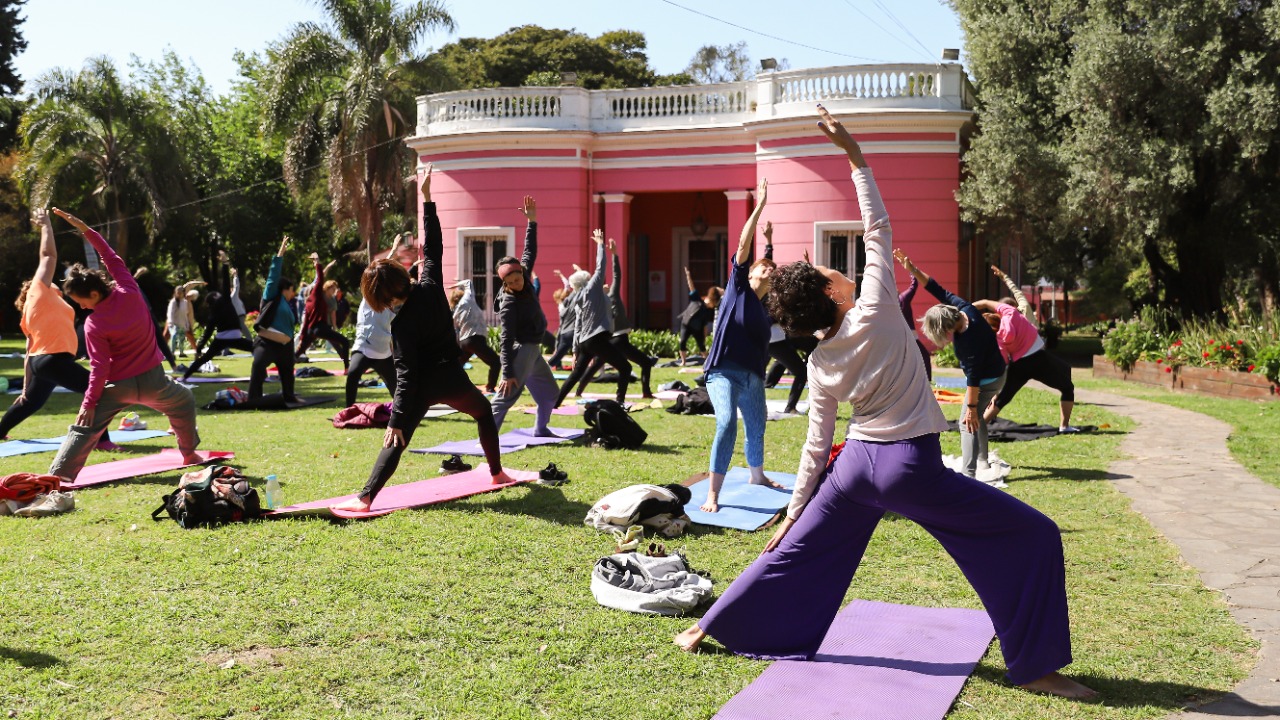 Grupo de personas realizando una clase de yoga al aire libre en el jardín de la Quinta Trabucco, con un edificio de color rosa de fondo.